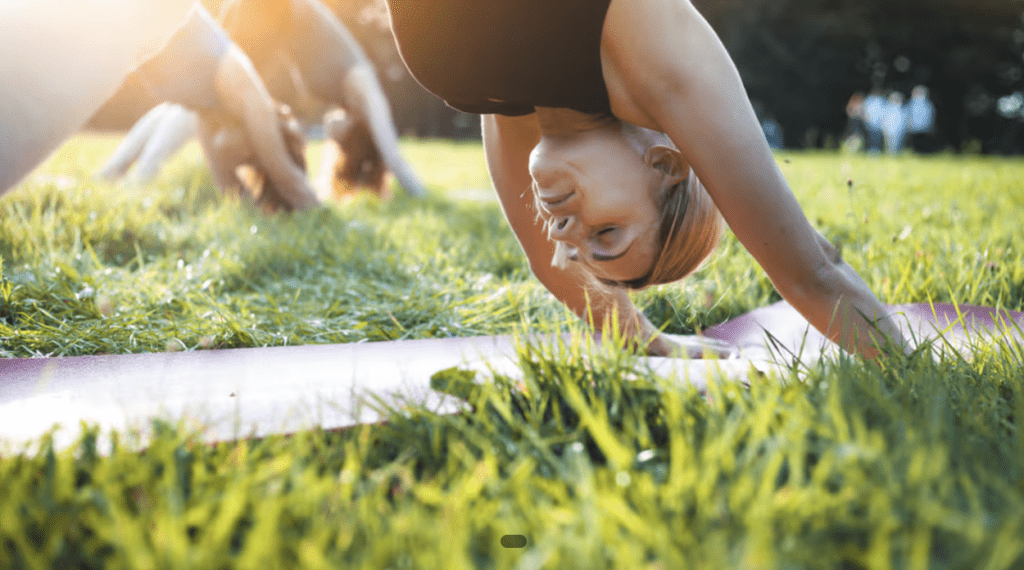 A group of people practice yoga outdoors on grass, with a woman in the foreground holding a downward-facing dog pose on a yoga mat in the sunlight.