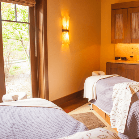Two massage tables with light purple blankets are set up in a cozy, softly lit spa room with a large window, warm beige walls, and wooden accents. Towels and robes are neatly placed on each table.