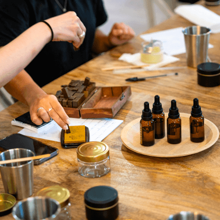 Two people sit at a wooden table with amber dropper bottles, jars, chocolate pieces, papers, and pens. One person points at a phone while the other handles chocolate.