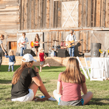 Two girls sit on grass watching a live band perform on an outdoor stage in front of a rustic wooden building. Children play nearby, and a table with white chairs is set up to the right. The scene is bright and sunny.