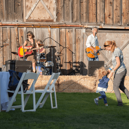 A band performs on a stage made of hay bales in front of a wooden barn while a woman dances with a small child on the grass. White folding chairs and a table are visible in the foreground.