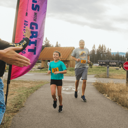 A young girl and an adult man, both wearing race bibs, run on a paved path toward a finish line. The girl smiles as she approaches a cheering hand for a high five. Trees and buildings are visible in the background.
