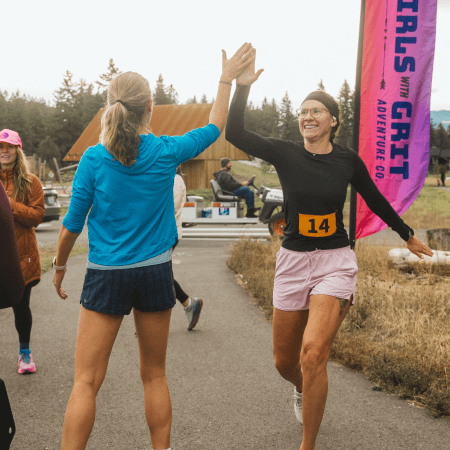 Two women high-five each other at an outdoor running event. One wears a race bib number 14 and shorts, the other wears a blue jacket. People and a “Girls With Grit” banner are in the background.