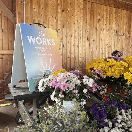 A display of colorful flowers in buckets sits beside a sign reading “the WORKS: Hands-on Classes, DIY Kits, Corporate Events,” against a wooden wall in a rustic setting.