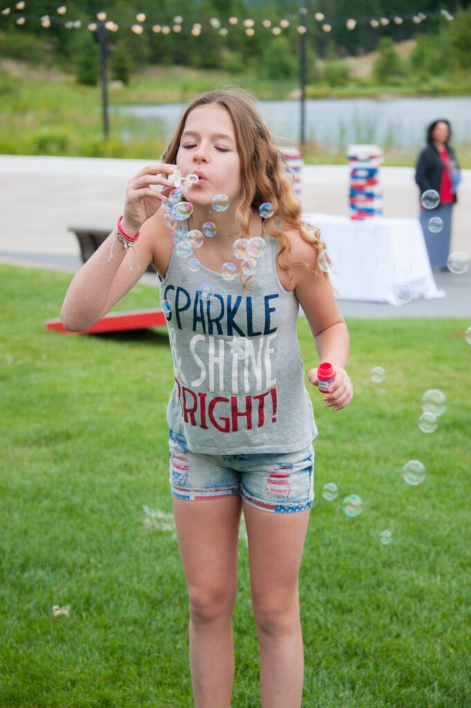 A young girl with long hair blows bubbles outside on a grassy lawn. She is wearing a grey tank top that says SPARKLE & SHINE BRIGHT! and denim shorts. There are scattered bubbles and blurred people in the background.