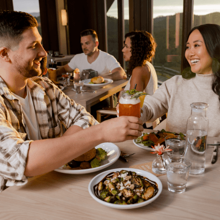 Two people sit at a restaurant table, smiling and clinking drinks. Their plates are filled with food, and another couple is seated at a table in the background by large windows with a scenic outdoor view.