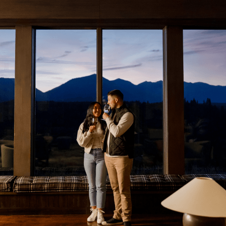 A couple stands by large windows at dusk, holding glasses and smiling at each other. Mountains and a darkening sky are visible outside. The indoor lighting is warm and cozy.