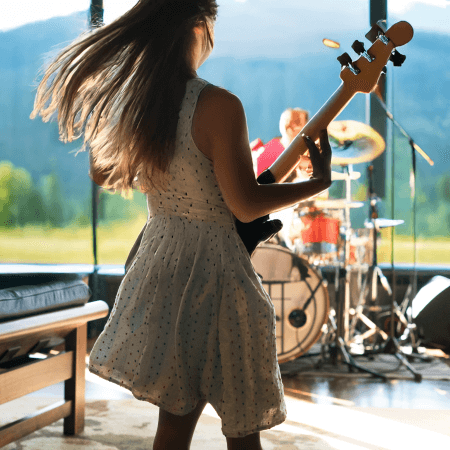 A woman in a white dress plays bass guitar onstage in front of large windows with mountains in the background, while a drummer performs in the distance. Natural light fills the space.