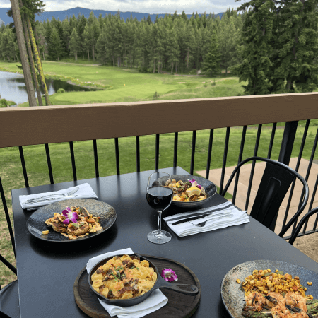 A table set with three plates of pasta, a glass of red wine, and napkins overlooks a lush golf course and pine trees, with mountains visible in the distance.