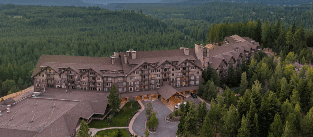 A large lodge-style hotel surrounded by dense green forest, with mountains visible in the background under a cloudy sky. The building features brown rooftops and multiple windows.