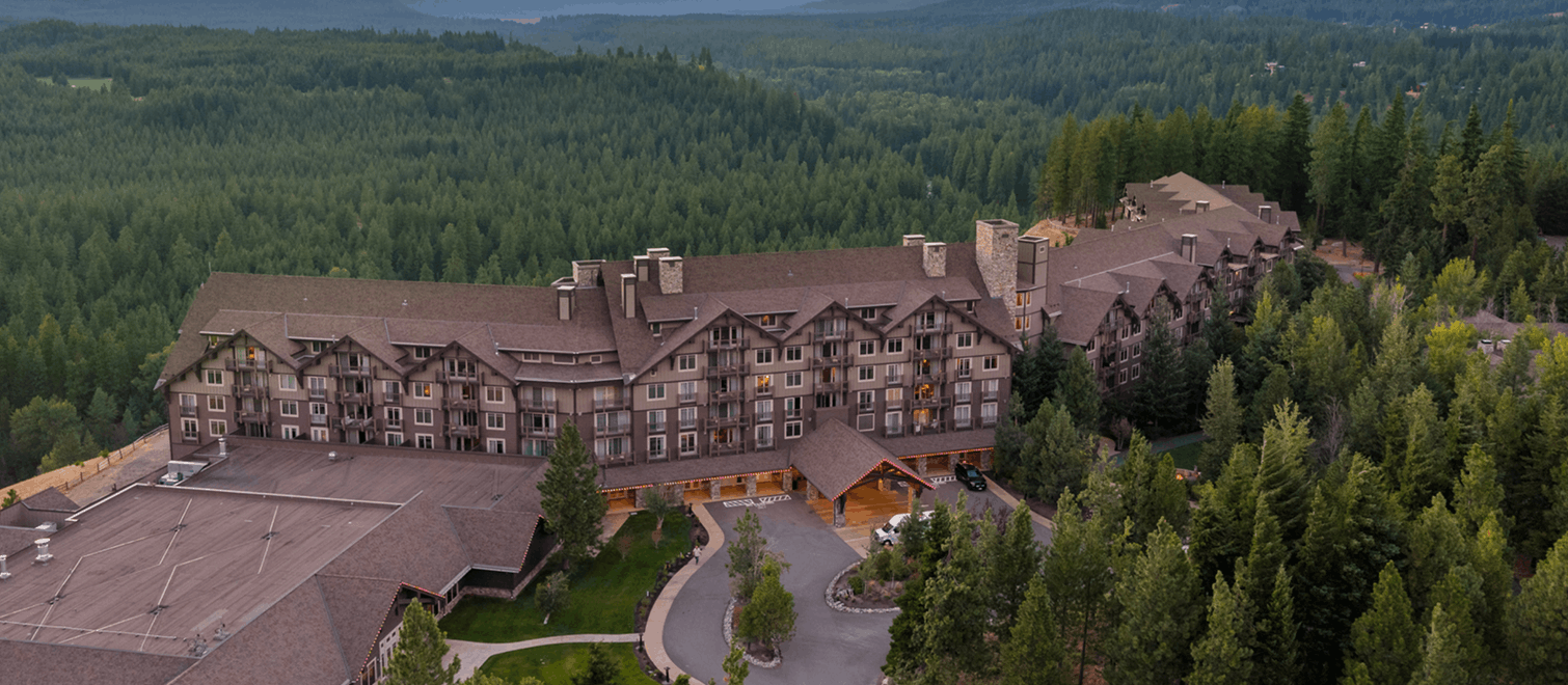 A large lodge-style hotel surrounded by dense green forest, with mountains visible in the background under a cloudy sky. The building features brown rooftops and multiple windows.