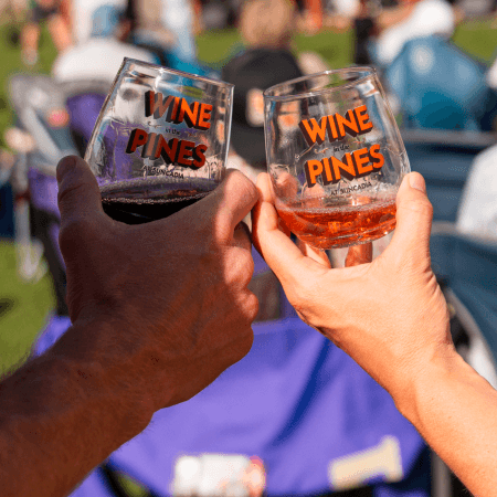 Two people clink glasses labeled Wine in the Pines at an outdoor event, with a blurred background of people sitting on chairs and grass, suggesting a festival atmosphere.