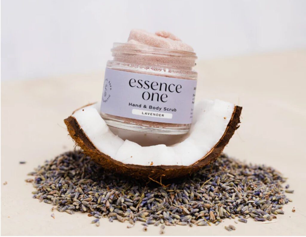 A jar of Essence One hand and body scrub with lavender sits in a halved coconut, surrounded by dried lavender buds on a light surface.