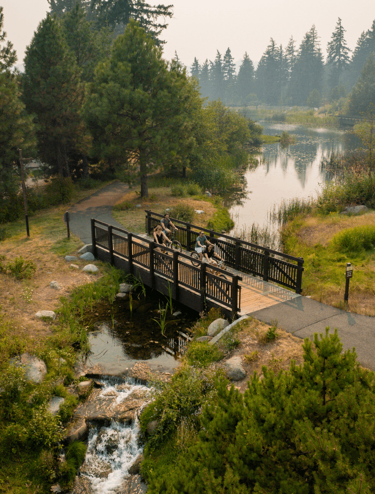 Aerial view of three people with bicycles standing on a wooden bridge over a creek, surrounded by trees, grass, and a pond in a natural park setting—perfect for experiencing Spring events at Suncadia.