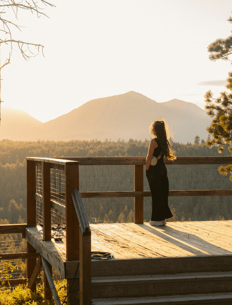 A woman in a long black dress stands on a wooden deck, gazing at forested mountains bathed in golden sunset light—an idyllic backdrop for Spring events at Suncadia, with trees and hazy peaks stretching under a clear sky.