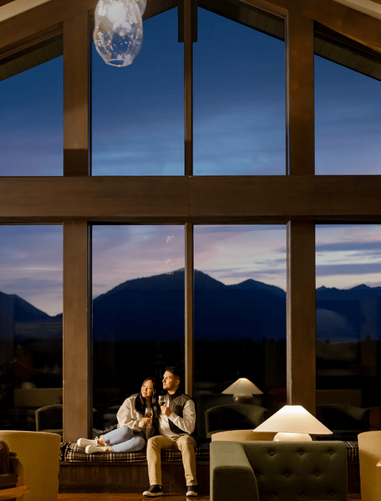 A couple sits on a window seat inside a cozy room, holding drinks and gazing at each other, with large windows framing a beautiful mountain view at dusk—perfect for relaxing after enjoying spring events at Suncadia. A lamp and green chair add warmth.