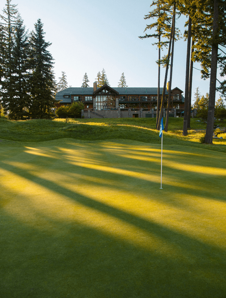 A golf green with a flagstick in the foreground is lit by late afternoon sunlight. In the background, a large lodge-style building—host to Spring events at Suncadia—is surrounded by tall pine trees under a clear sky.