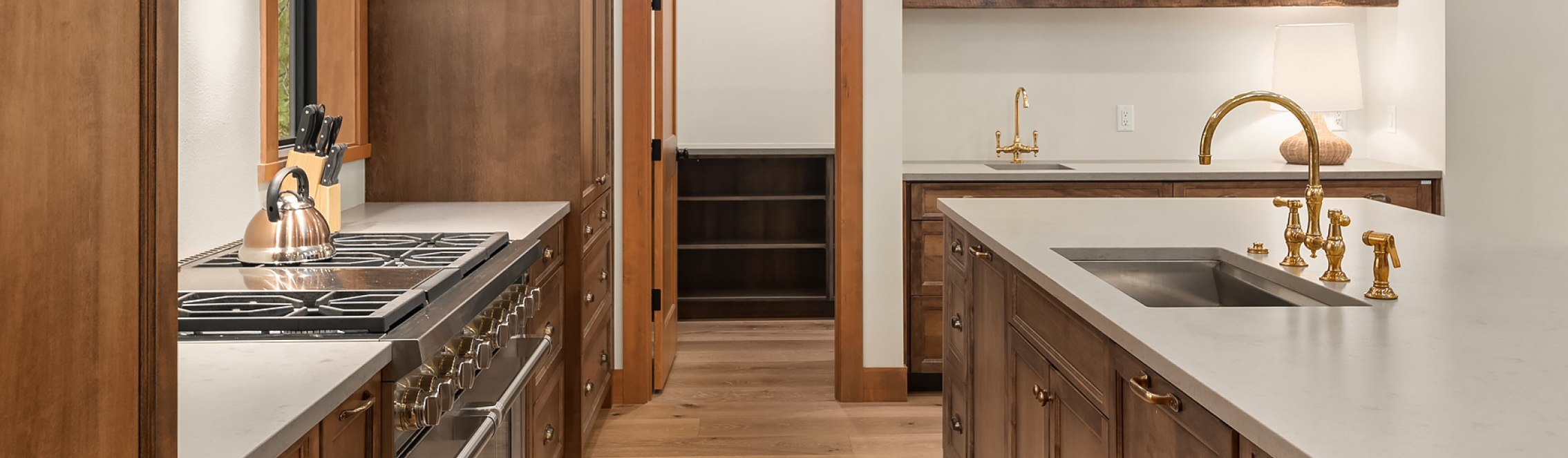 Modern kitchen with wooden cabinets, white countertops, gold fixtures, a stainless steel stove with a kettle, a sink on an island, knife block, lamp, and open pantry visible in the background.