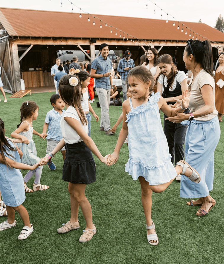 Children and adults joyfully dance and hold hands outdoors on green grass, surrounded by string lights and rustic wooden buildings on a sunny day.
