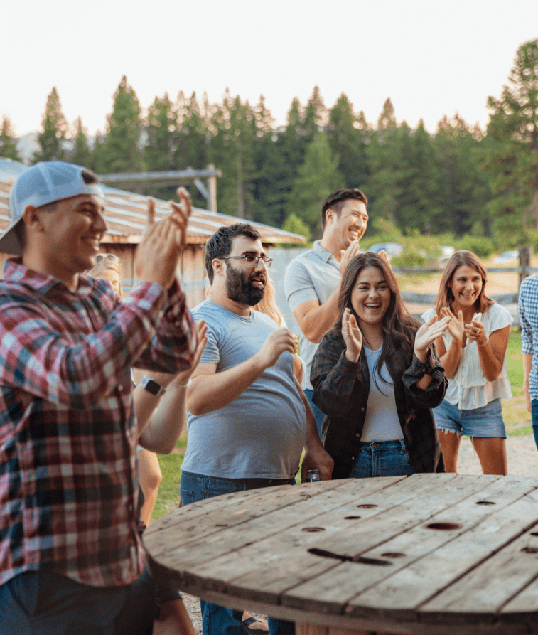 A group of young adults stands outdoors, smiling and clapping near a wooden spool table. Trees and buildings are in the background, suggesting a casual, social gathering in a rustic setting.