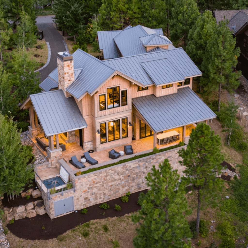 Aerial view of a modern wooden house with large windows, a stone patio, outdoor loungers, and a small pool, surrounded by trees and greenery in a forested area.