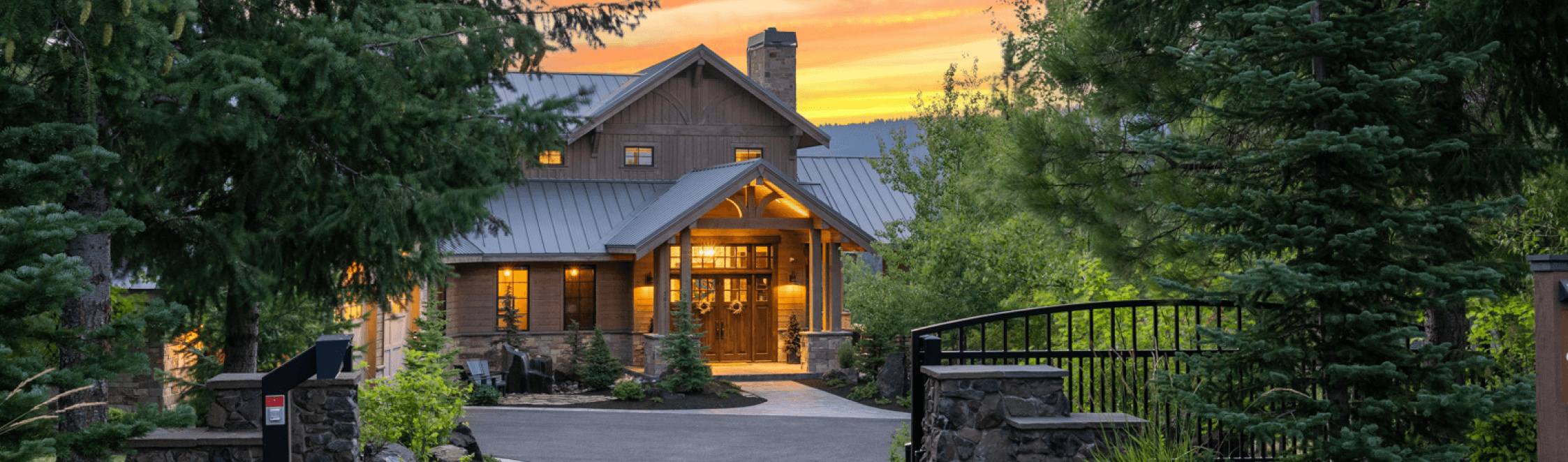 A large, rustic house with warm lights shines through windows at sunset, surrounded by lush trees and greenery, with a gated driveway leading to the entrance.