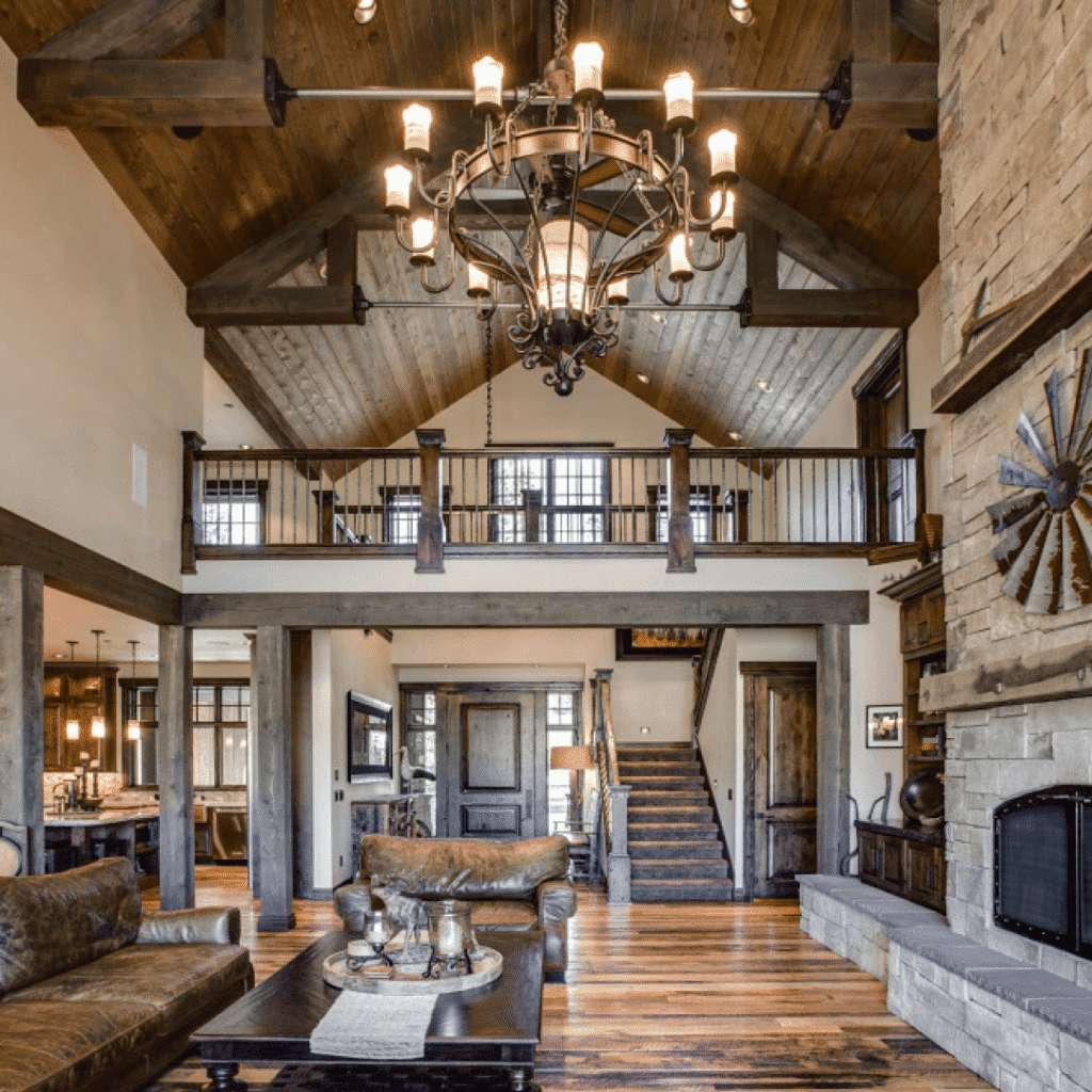 A rustic living room with exposed wooden beams, a large stone fireplace, leather furniture, a chandelier, and an open loft area above with a wooden staircase and railing.