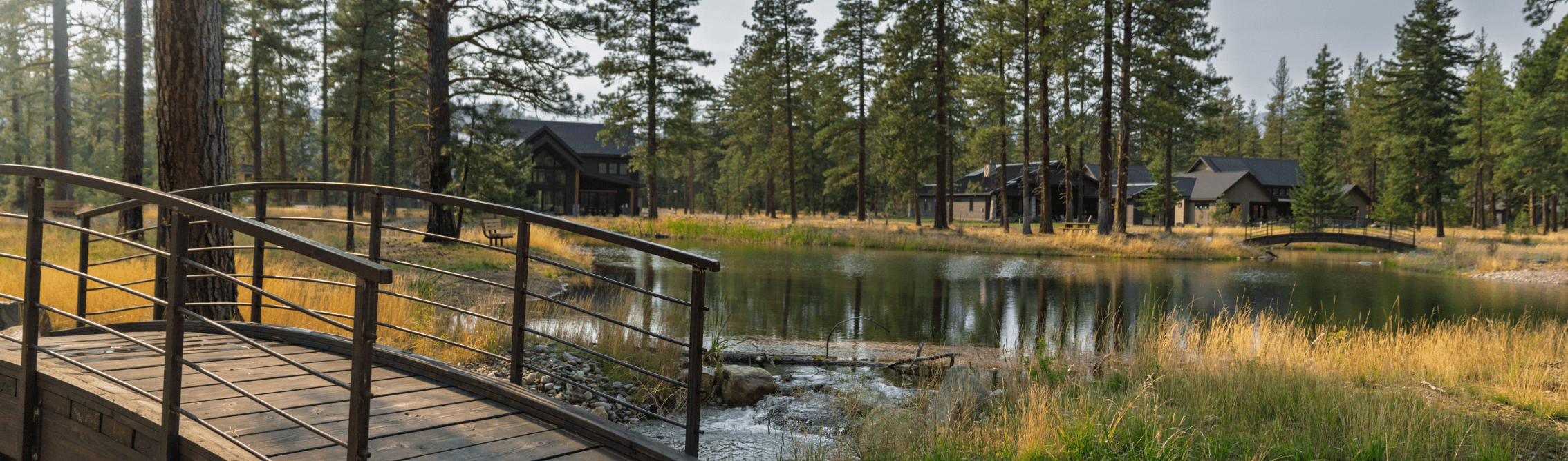 A wooden footbridge crosses a small stream leading to a pond with tall trees and several dark-roofed cabins in the background, surrounded by grass and forest—perfect scenery for the Suncadia Real Estate Brokerage Team to showcase.