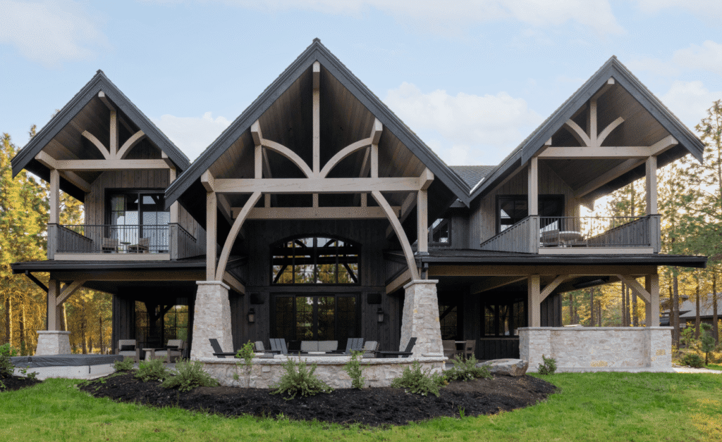 A modern rustic house with steep pitched roofs, large wooden beams, stone columns, upper balconies, floor-to-ceiling windows, and a spacious patio, surrounded by greenery and trees under a blue sky.