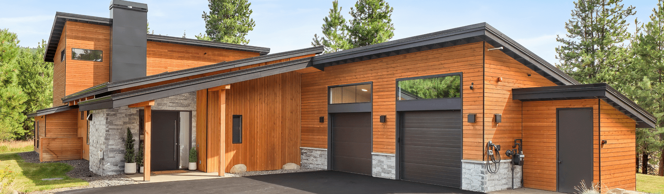 Modern house with a combination of wood and stone exterior, large windows, two garage doors, and dark sloped roofs, surrounded by green trees and a clear sky.