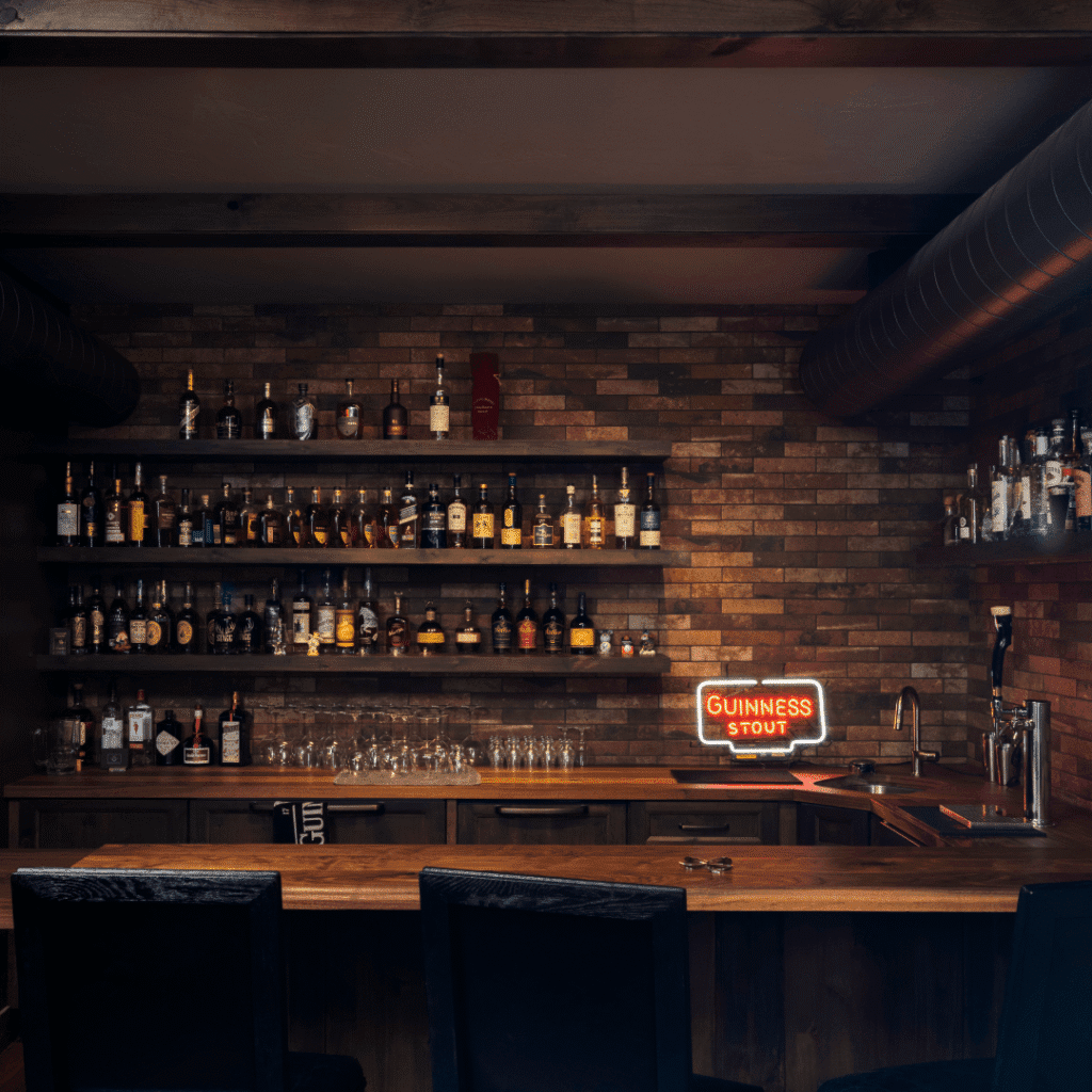 A cozy home bar with wooden shelves stocked with various liquor bottles, glassware beneath, a wooden counter, and a neon Guinness Stout sign glowing on a brick wall in the background.