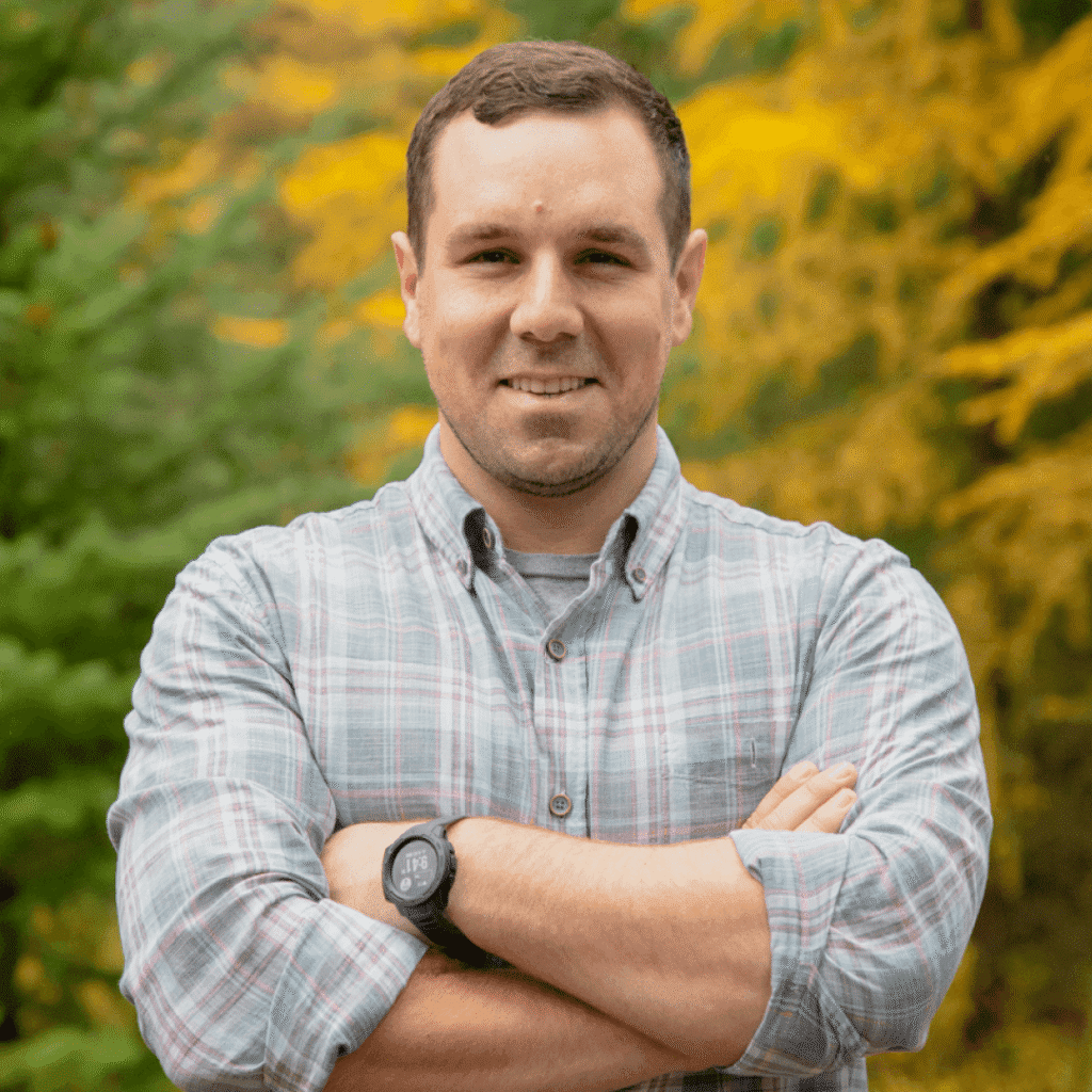 A man with short brown hair, wearing a plaid shirt and a watch, stands outdoors with arms crossed and smiles. Green and yellow foliage in the background suggests a fall setting.