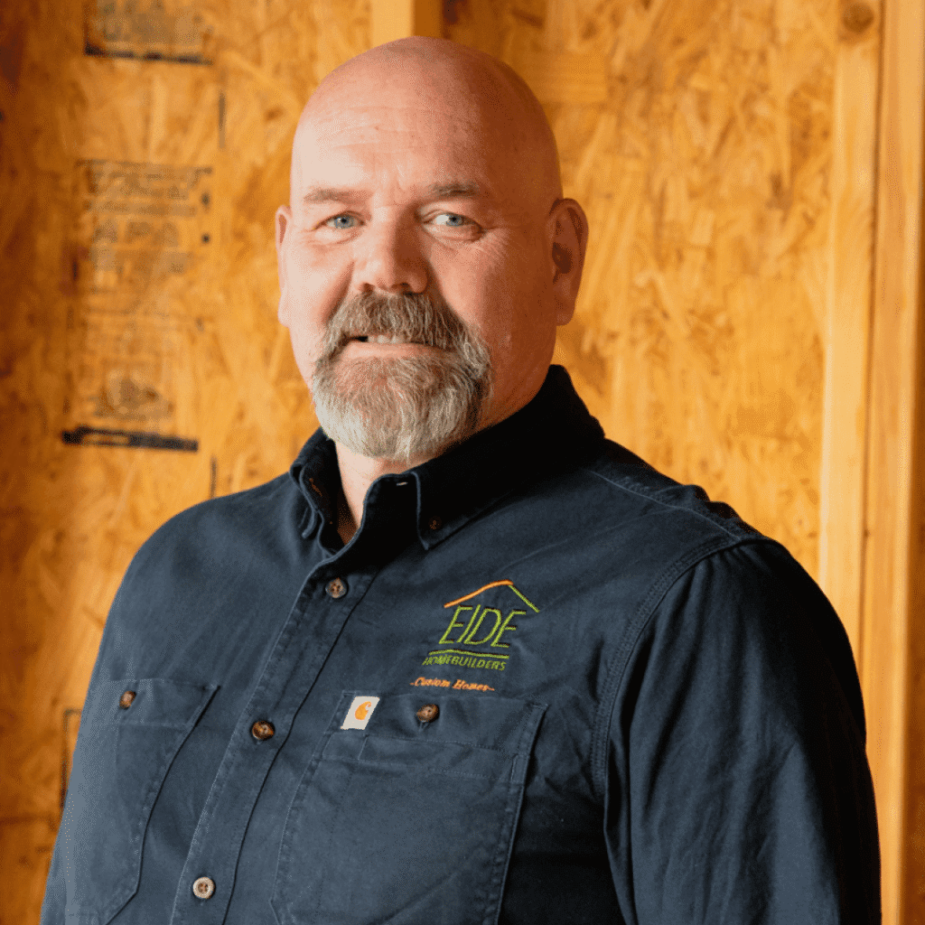 A man with a bald head and gray mustache and beard stands indoors wearing a dark blue button-up shirt with the EIDE logo and text, against a background of unfinished wooden walls.