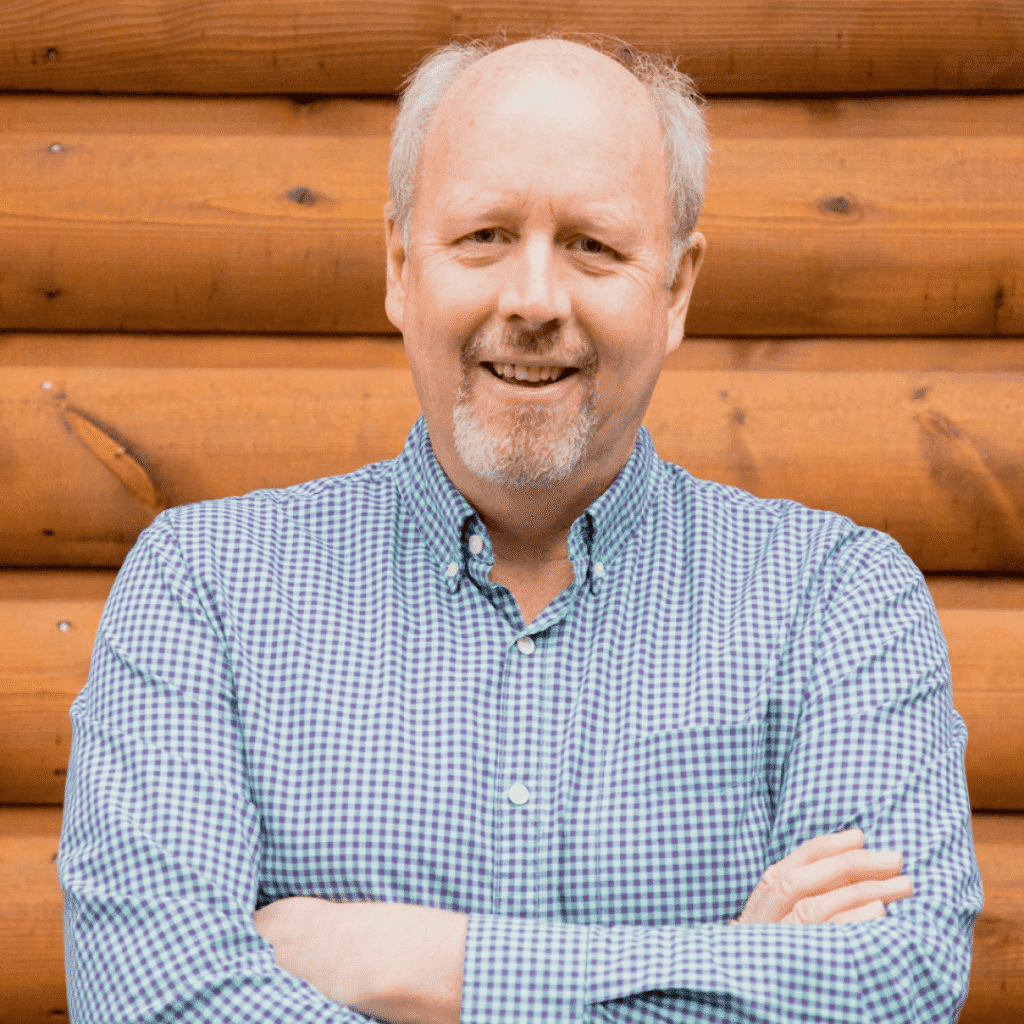 A middle-aged man with short light hair and a beard, wearing a blue and white checkered shirt, stands smiling with arms crossed in front of a wooden log wall.