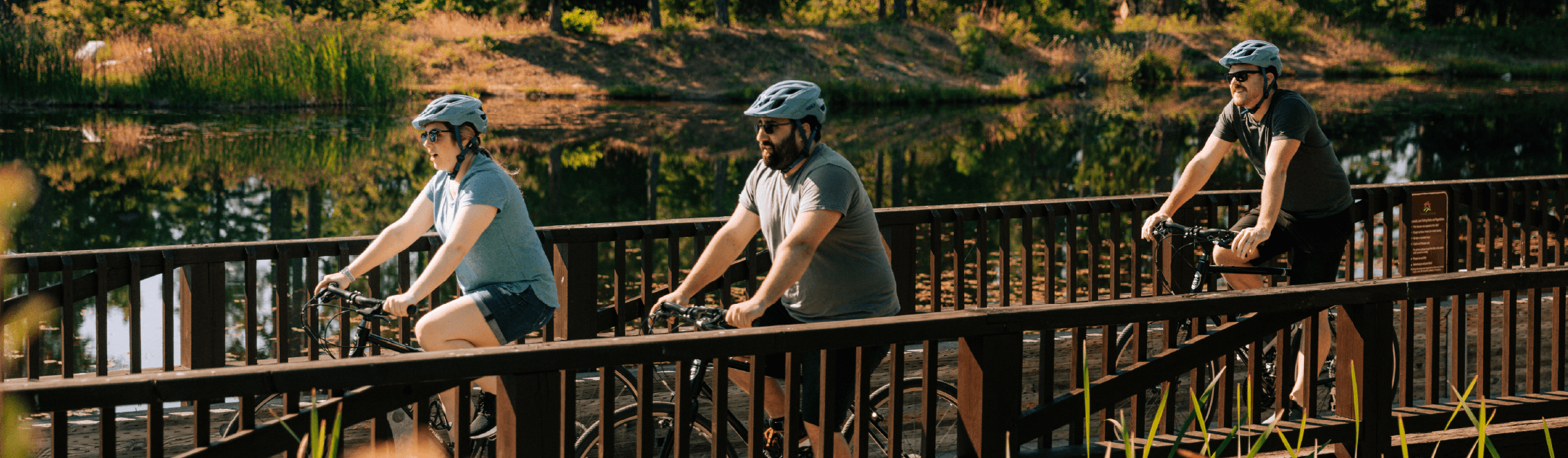 Three people wearing helmets ride bicycles across a wooden bridge beside a calm, tree-lined body of water on a sunny day.