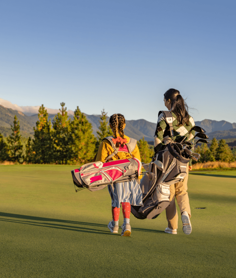 A woman and a young girl, both carrying golf bags, walk together across a green golf course with mountains and trees in the background under a clear blue sky.