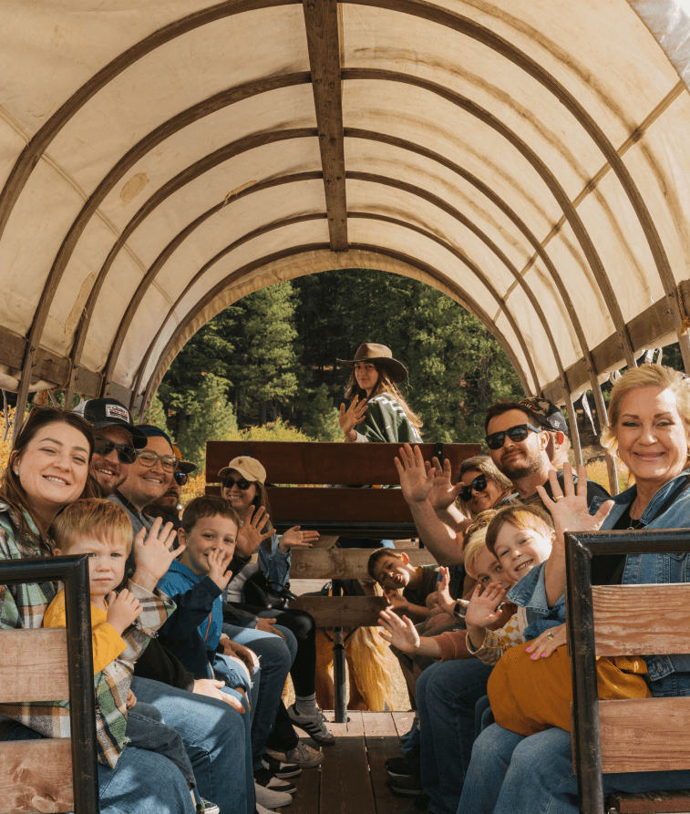 A group of people, including adults and children, smile and wave while sitting on benches inside a covered wagon. Trees and greenery are visible in the background. A person in a cowboy hat is seated at the front, holding reins.