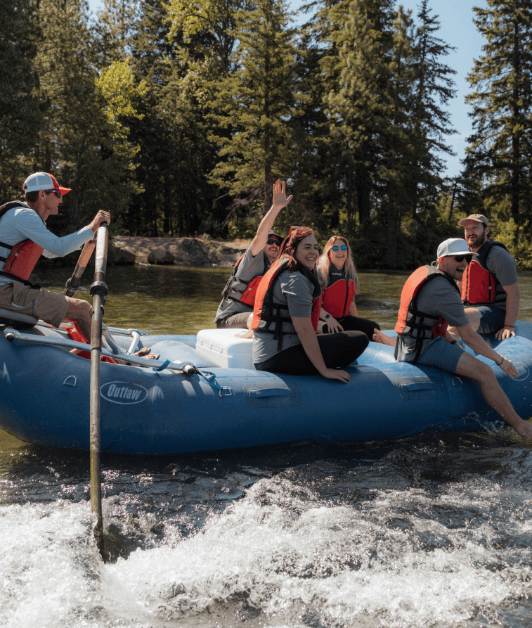 A group of six people wearing life jackets sit and smile on a blue inflatable raft as they paddle down a river surrounded by trees on a sunny day. One person waves to the camera.