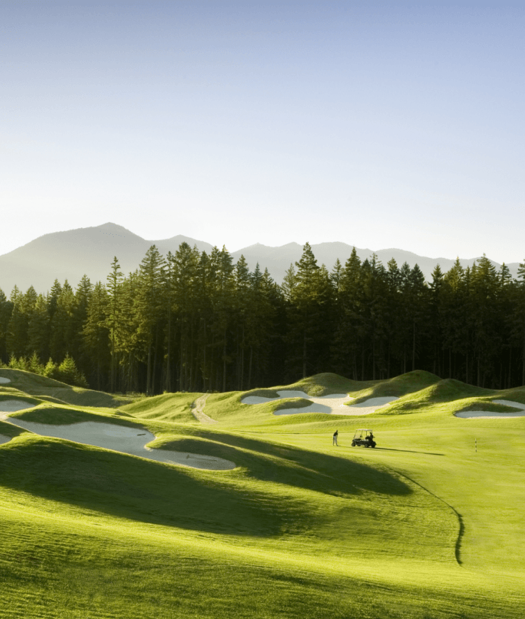 A lush green golf course with sand bunkers, two people near a golf cart, and tall pine trees in the background. Sunlight illuminates the rolling hills and distant mountains under a clear blue sky.