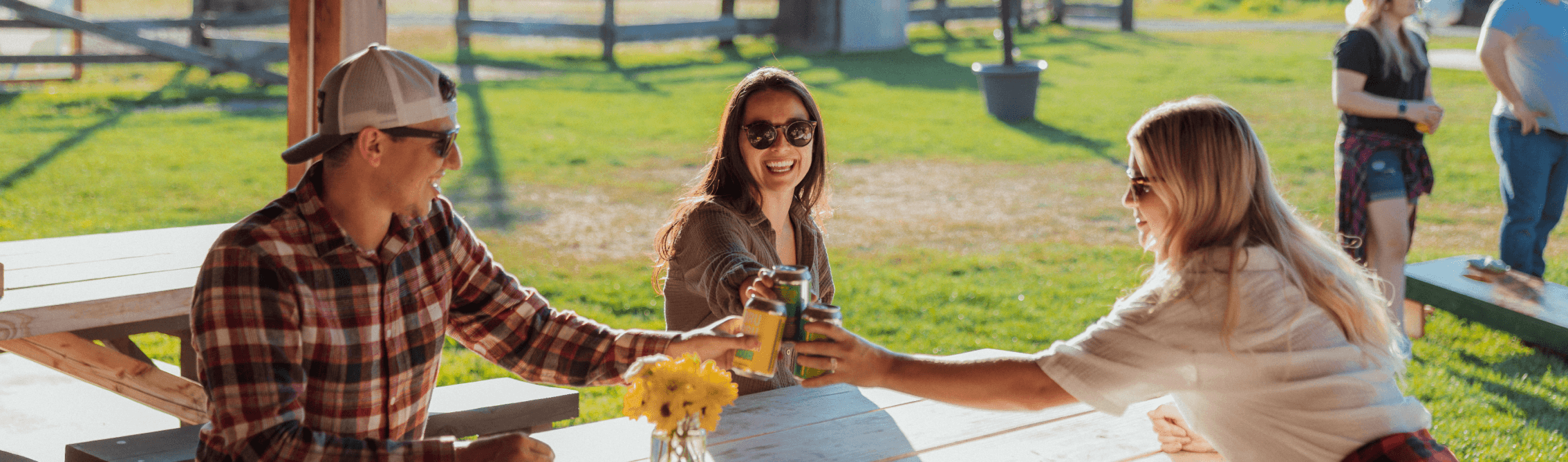 Three people sit at a picnic table outdoors, smiling and clinking drinks together. They are wearing sunglasses and casual clothes, enjoying a sunny day. Two other people are visible in the background.