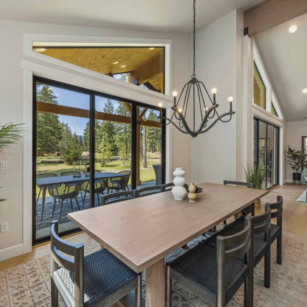 A modern dining room with a light wooden table, six dark chairs, and a black chandelier. Large windows reveal a patio with outdoor seating and a scenic green yard with trees. Natural light fills the space.