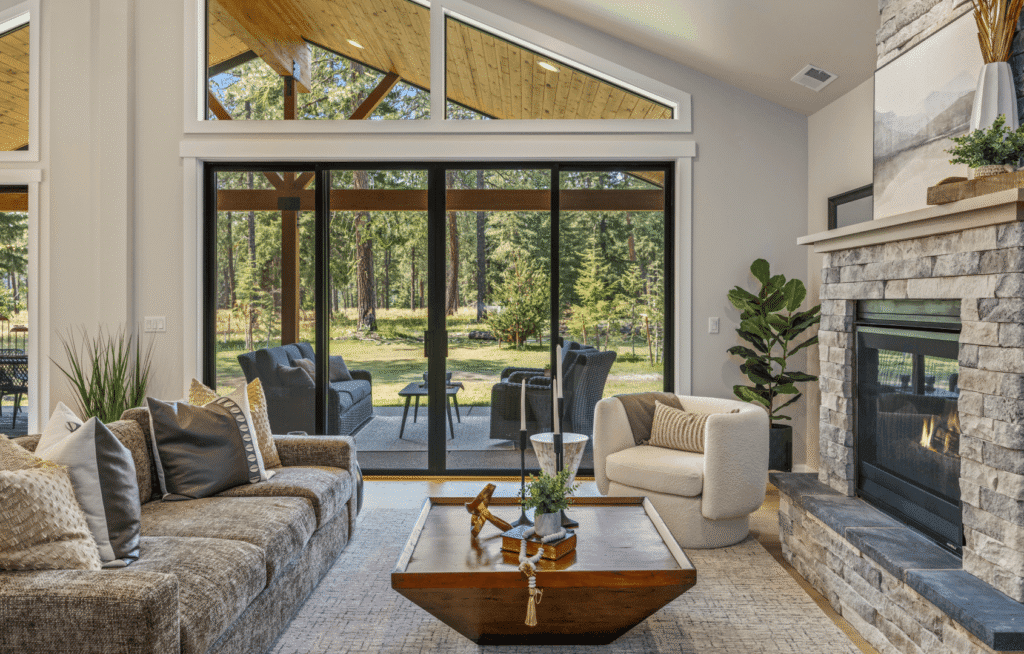 Modern living room with a stone fireplace, cozy seating, and a wooden coffee table. Large glass doors open to a patio with outdoor furniture, offering a scenic view of trees and greenery.