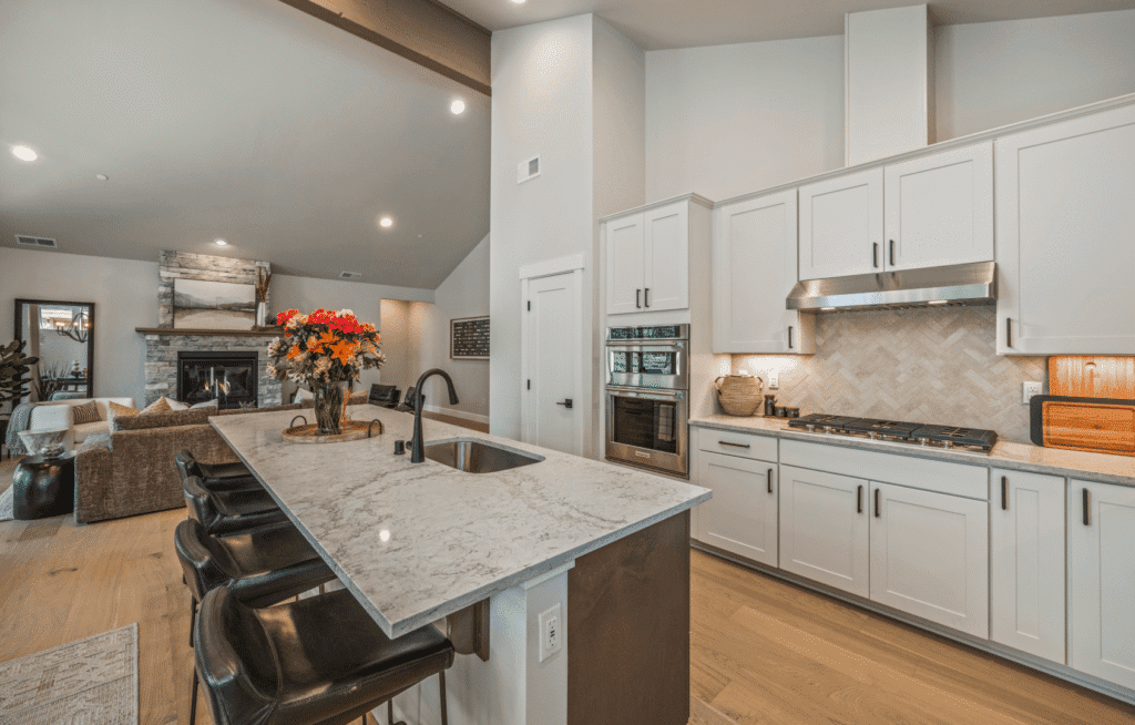 Modern kitchen with white cabinets, stainless steel appliances, a marble island with black barstools, and a vase of flowers; open layout connects to a cozy living area with a fireplace in the background.