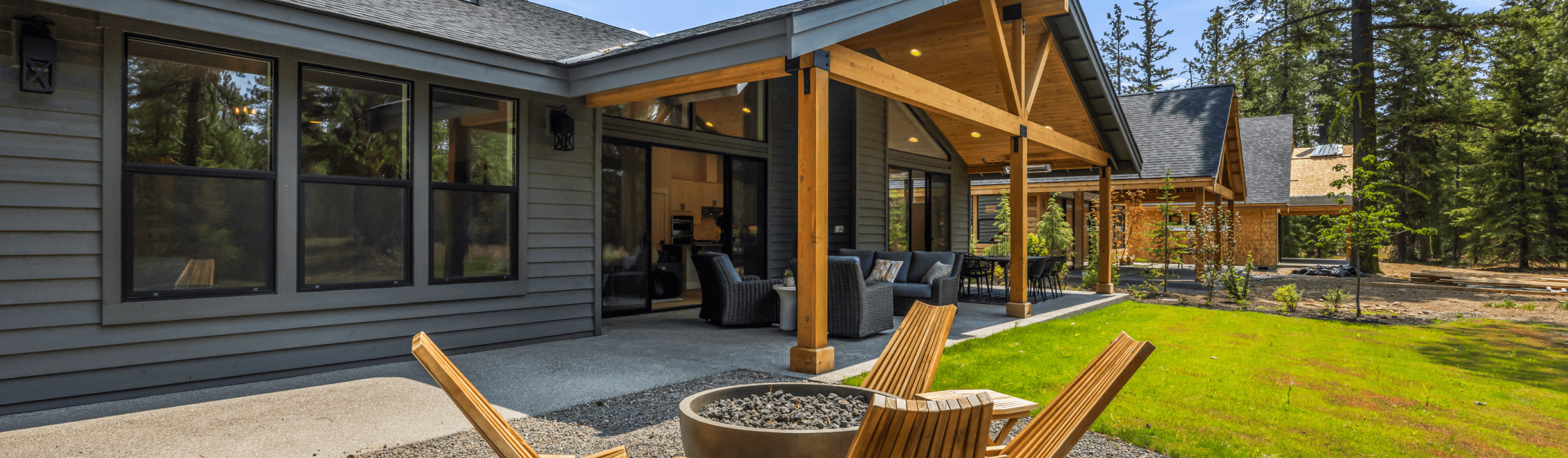 A modern house with gray siding and large windows features a covered patio with outdoor seating. In the foreground, wooden chairs surround a fire pit on a grassy lawn with trees in the background.