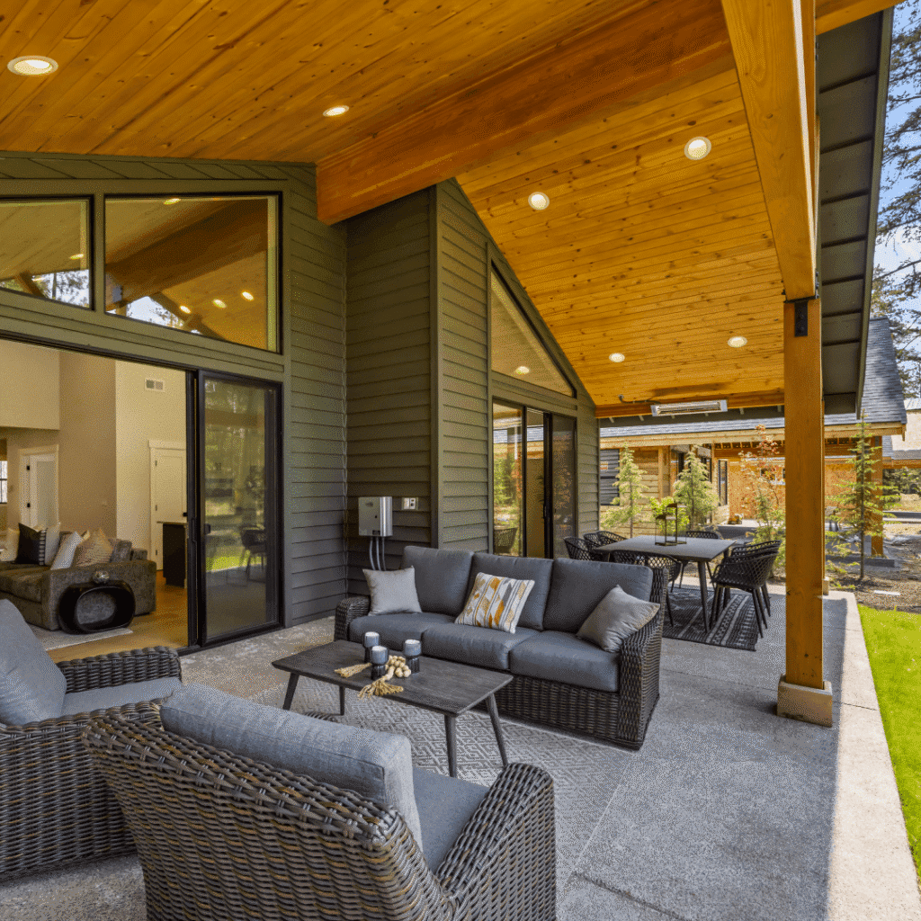 Modern covered patio with gray wicker furniture, gray cushions, and a wooden ceiling. Large glass doors lead to the interior. Outdoor seating area overlooks a green lawn and trees. Warm lighting creates a cozy atmosphere.