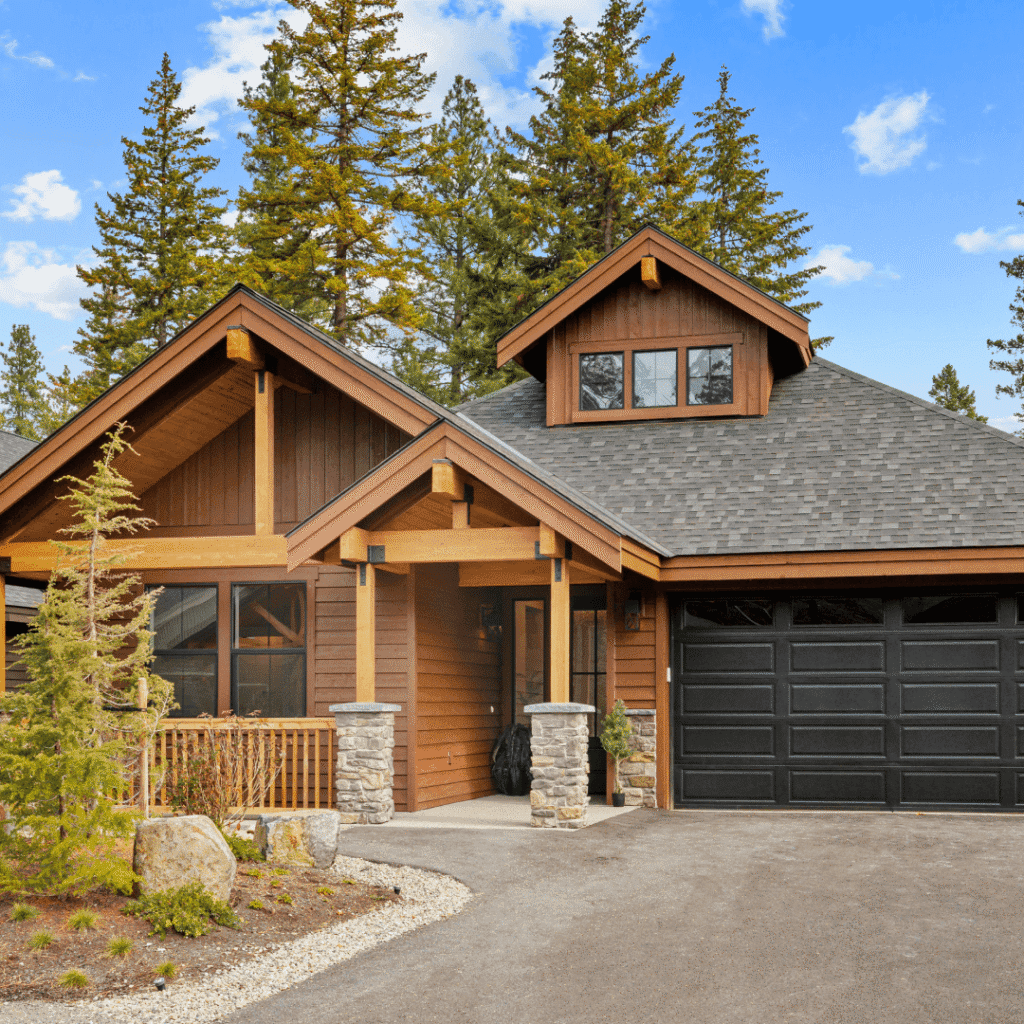 A modern cabin-style house with wood siding, stone pillars, black garage door, and surrounded by pine trees under a blue sky with scattered clouds.