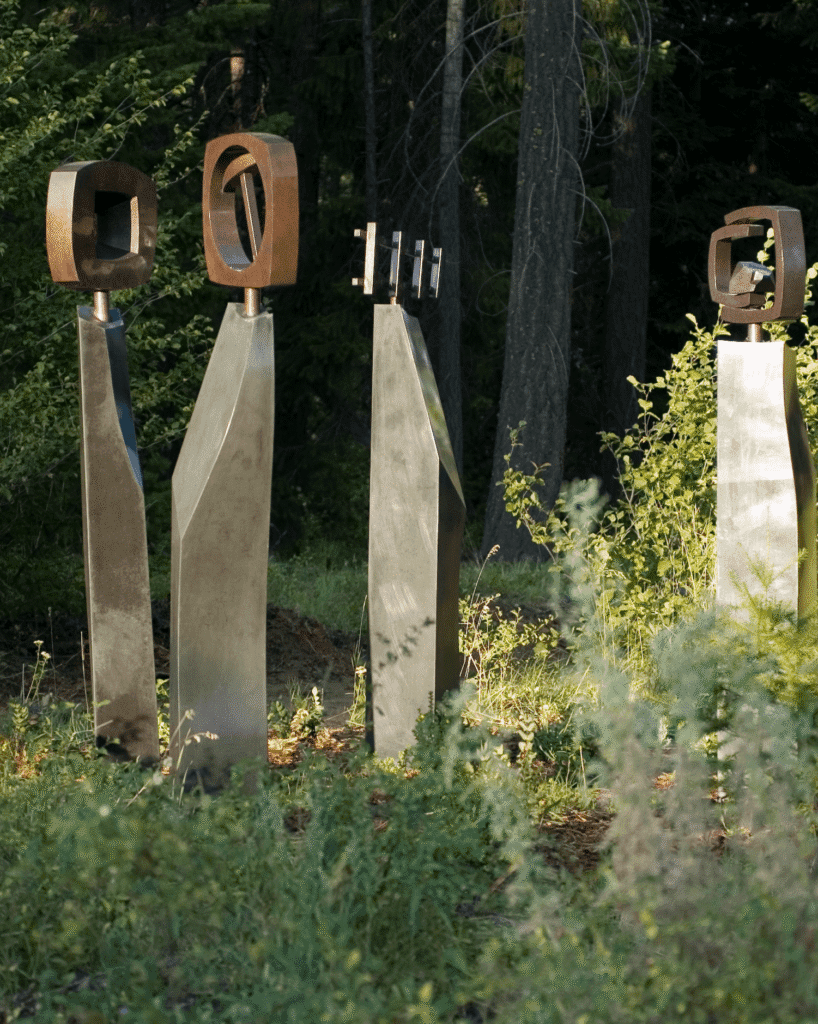 Three tall metal sculptures with abstract geometric shapes on top stand among green plants and trees in a forested area, with sunlight highlighting parts of the scene.