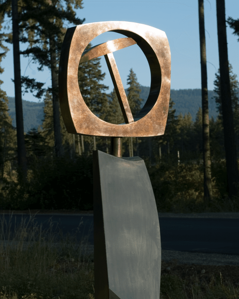 A modern outdoor sculpture featuring a bronze circular shape with an angled bar inside, mounted on a curved metal base, set against a backdrop of trees and mountains.