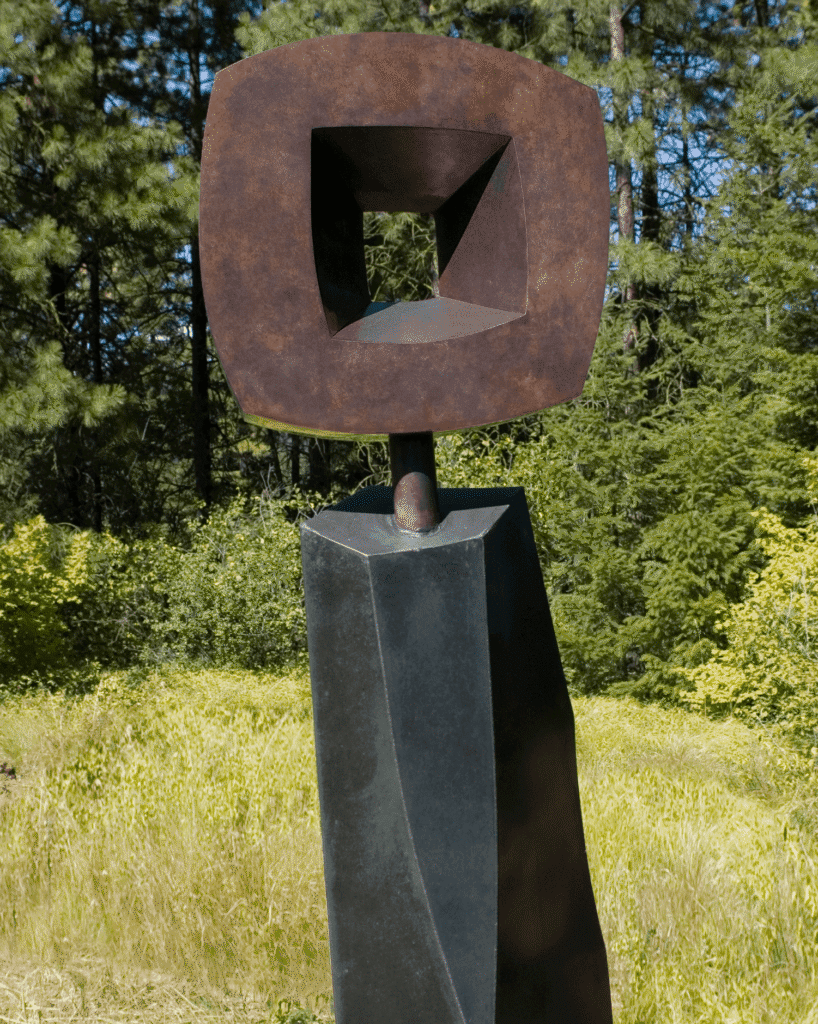 A modern abstract outdoor sculpture with a geometric, hollow brown square atop a tall, angular black base, set in a grassy area with trees in the background.