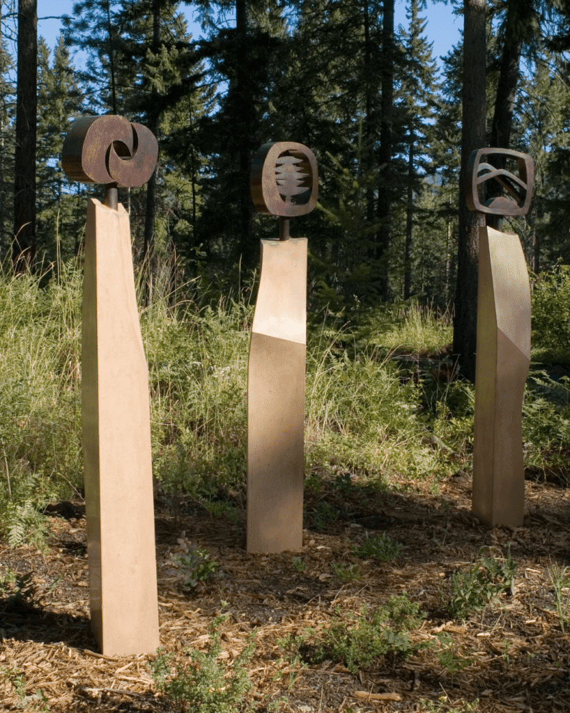 Three tall, angular stone pedestals stand in a forest clearing, each topped with a different abstract, circular metal sculpture against a backdrop of trees and greenery.